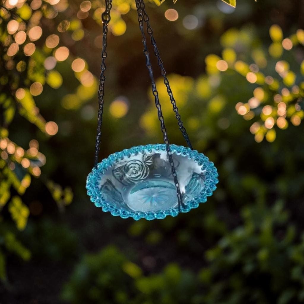 Gros plan sur un abreuvoir à oiseaux suspendu en verre turquoise à motif rose et bord perlé, chaînes de suspension, sur fond de jardin lumineux
