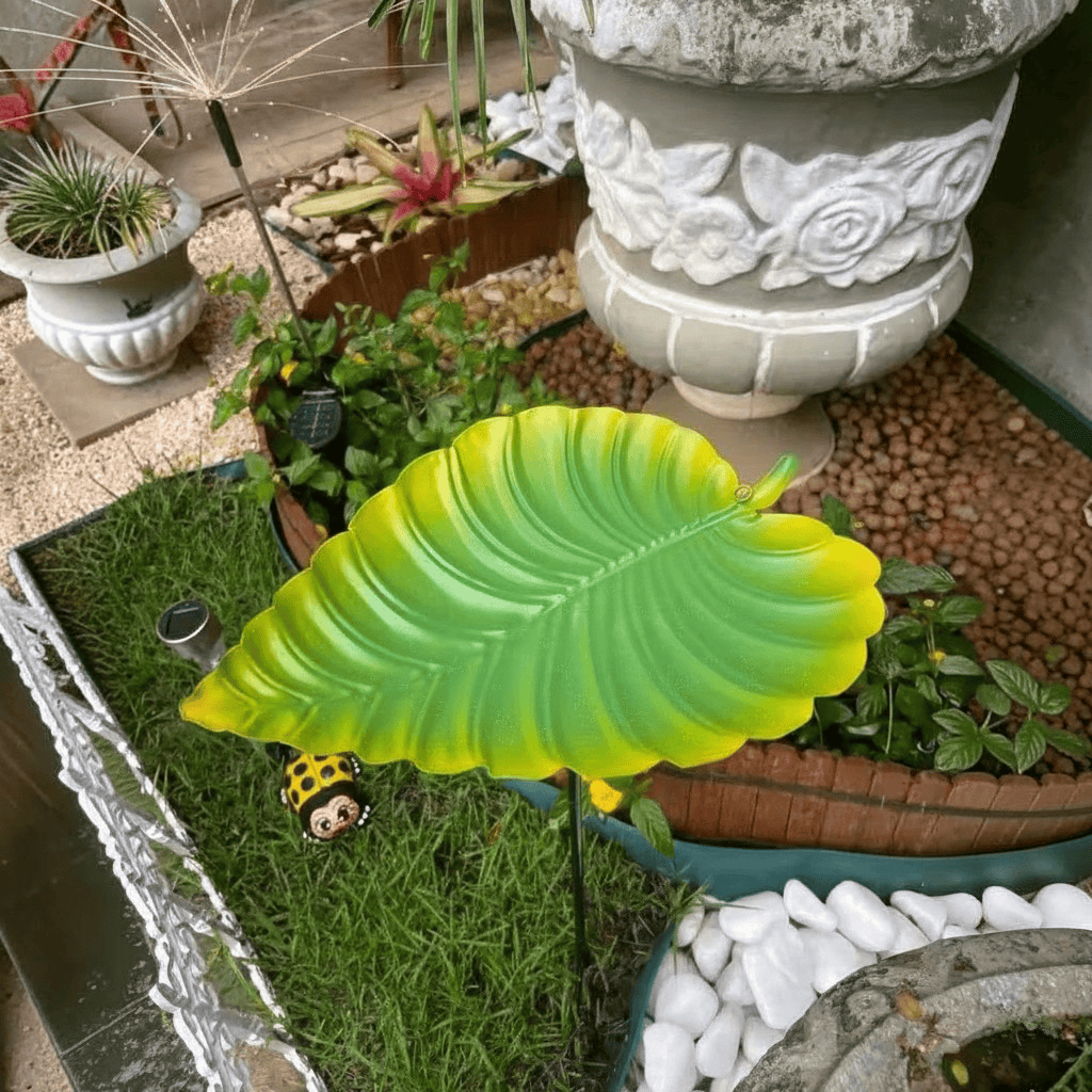 Abreuvoir à oiseaux en forme de feuille verte sur piquet, planté dans une jardinière fleurie sur une terrasse décorée de pots et galets

