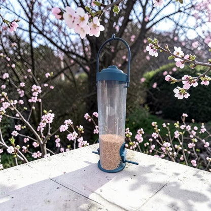 Mangeoire à oiseaux tubulaire grise remplie de graines posée sur une table en pierre, devant un cerisier en fleurs roses au printemps