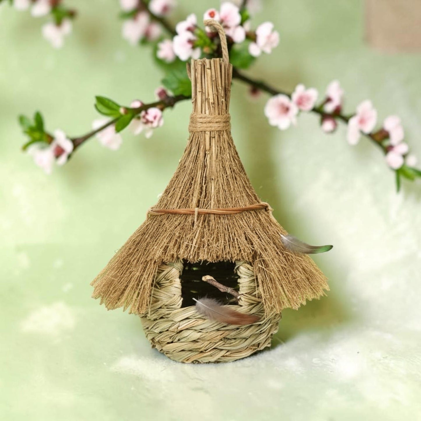 Nichoir en paille tressée toit de chaume suspendu à une branche de cerisier en fleurs roses, décoré de petites plumes, sur fond vert doux