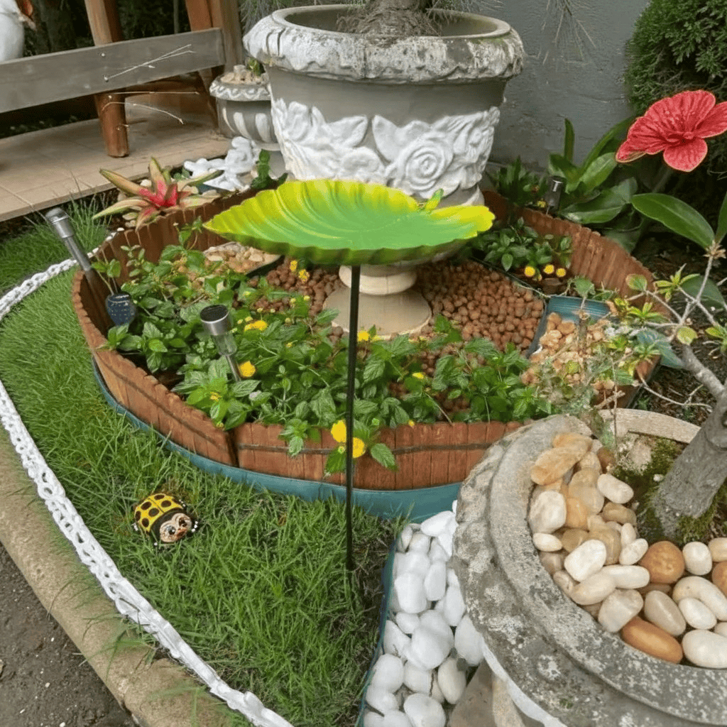 Abreuvoir à oiseaux feuille verte sur piquet installé au centre d'un massif de fleurs et plantes, avec vasque en pierre et fontaine de jardin