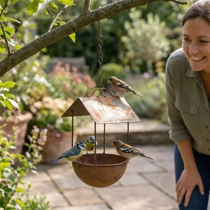 Mésange bleue et pinsons posés sur un abreuvoir en fer rouillé à toit, dans un jardin fleuri sous le regard d'une femme souriante